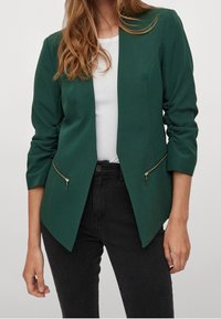 Grön skräddarsydd kavaj med en smal design, som har silverfärgade dragkedjefickor och trekvartsärmar, tillverkad av ett mjukt tyg.