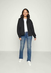 Sweatshirt preto com fecho, ombros caídos, t-shirt branca com gola redonda, jeans claros de boca de sino e sapatilhas brancas. Fundo simples.