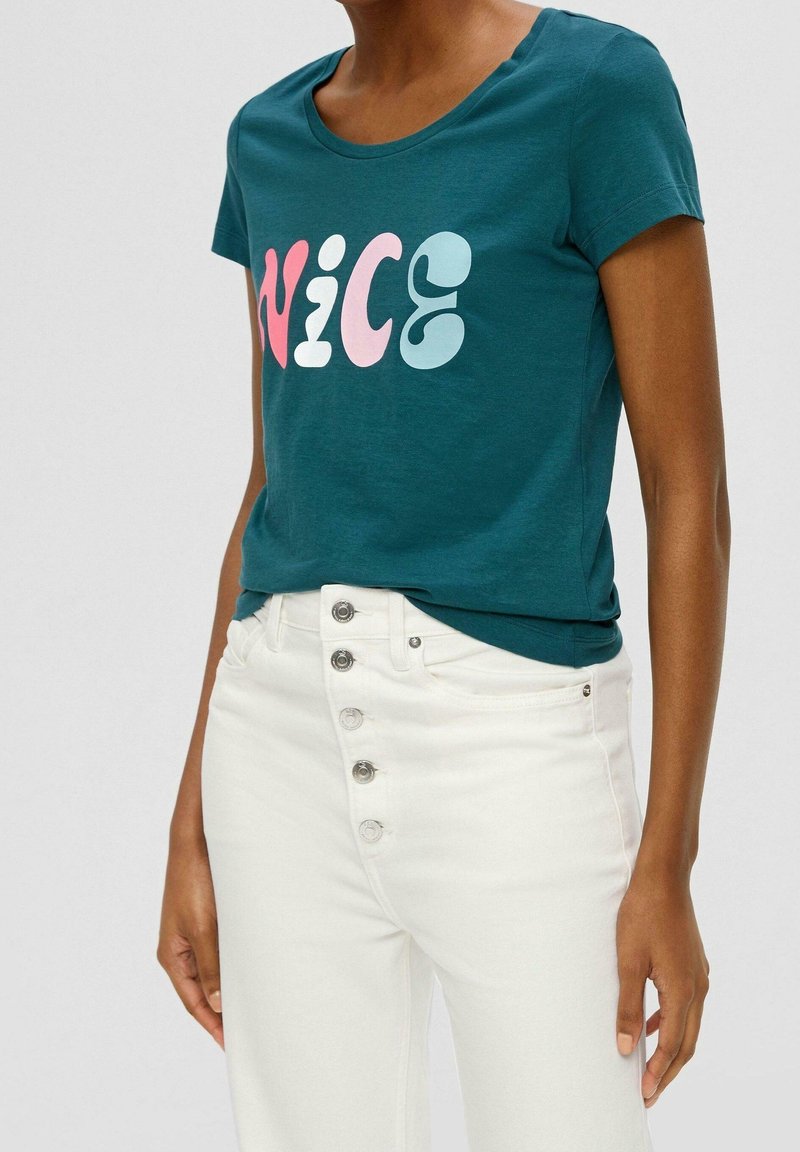 Personne portant un t-shirt turquoise à manches courtes avec "NICE" en lettres colorées et un pantalon blanc taille haute avec cinq boutons visibles.