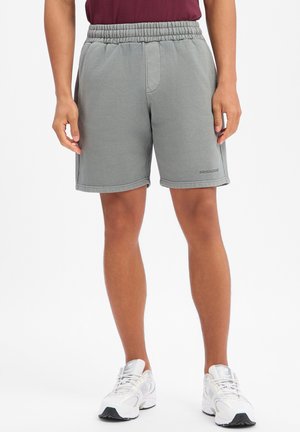 Man draagt grijze shorts met elastische taille en witte sneakers, staat met ontspannen handen langs zijn zij op een witte achtergrond.