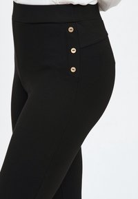 Zwarte legging gemaakt van rekbare stof, met een hoge tailleband en drie gouden knoopaccenten aan de zijkant voor extra detail.