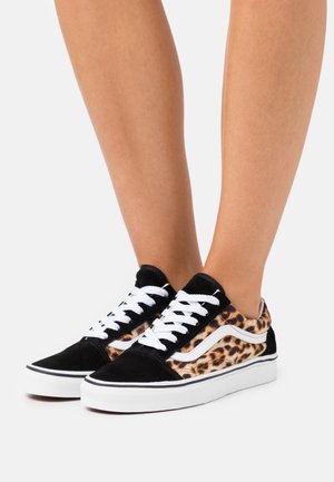 Benedenbenen met zwart-witte sneakers met zijkanten in luipaardprint en witte veters tegen een effen witte achtergrond.