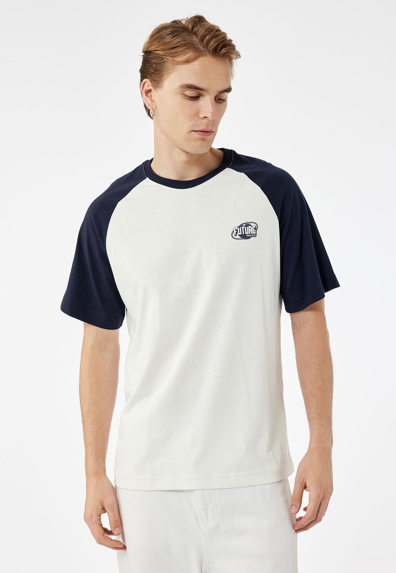 T-Shirt mit kurzen Ärmeln, cremefarbener Körper mit marineblauen Raglanärmeln, runder Ausschnitt, mit einem kleinen Grafiklogo auf der linken Brust. Aus weichem Baumwollmaterial.