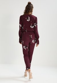 Mono floral burdeos con mangas largas, cintura elástica y piernas estrechas, con patrones de flores en crema y rosa. Tacones de aguja clásicos.