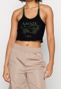 En person står klädd i en svart mesh-crop-top med gröna grafiker, kombinerat med ljusbeige cargobyxor, vilket visar en avslappnad modestil.