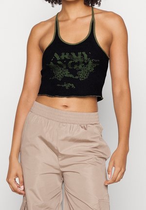Una persona está de pie vistiendo un top corto de malla negro con gráficos verdes, combinado con pantalones cargo beige claros, mostrando un estilo de moda casual.