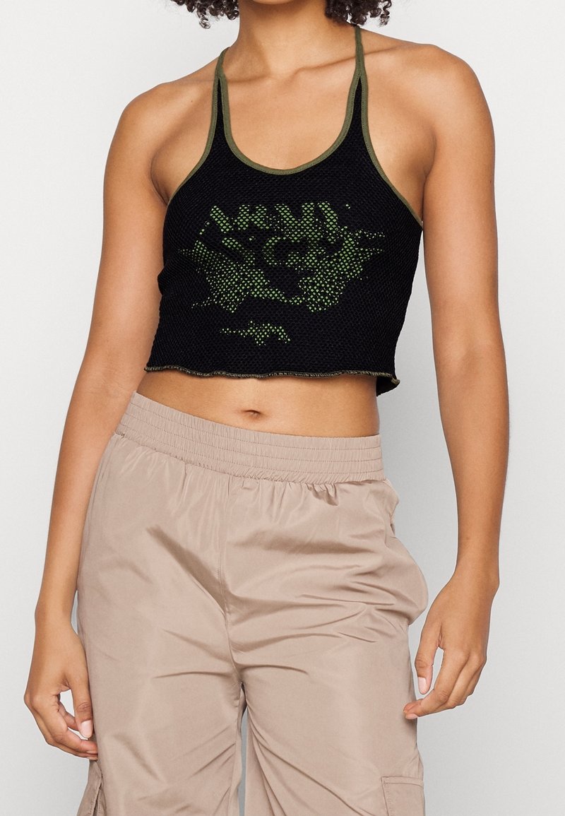 En person står klädd i en svart mesh-crop-top med gröna grafiker, kombinerat med ljusbeige cargobyxor, vilket visar en avslappnad modestil.
