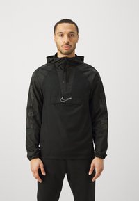 Man in een zwarte Nike hoodie met halve rits en logo aan de voorkant, staand met ontspannen armen tegen een effen lichte achtergrond.