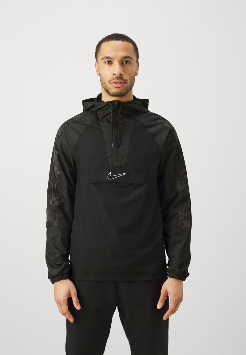 Man in een zwarte Nike hoodie met halve rits en logo aan de voorkant, staand met ontspannen armen tegen een effen lichte achtergrond.