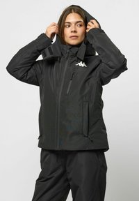 Schwarze wasserdichte Jacke mit einem Reißverschluss und einem Klettverschluss auf der Vorderseite, verstellbarer Kapuze und zwei Seitentaschen. Mit einem weißen Logo auf der Brust.