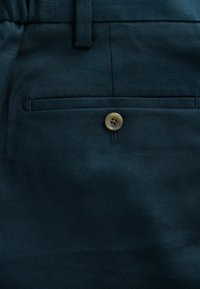 Tissu bleu foncé avec une poche arrière sécurisée par un seul bouton beige et une boucle de ceinture au-dessus de la poche.