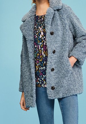 Kobieta w niebieskim, puszystym płaszczu zapinanym na guziki, na ciemnej kwiecistej bluzce i niebieskich dżinsach, z jedną ręką w kieszeni płaszcza na tle niebieskiego tła.