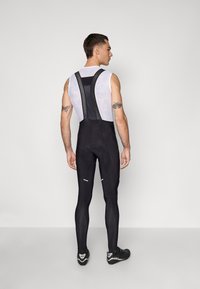 Schwarze Radlerbib-Shorts mit Mesh-Trägern, enganliegendem Design und reflektierenden Akzenten. Zeigt Tattoos auf sichtbarer Haut.