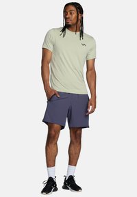 Camicetta verde chiaro a maniche corte con logo sul petto, abbinata a pantaloni corti blu scuro. Il modello è in piedi con una mano in tasca, indossando sneakers nere.
