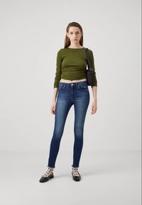 Top verde cropped de mangas longas combinado com jeans skinny azuis. Acessórios incluem uma pequena bolsa cruzada preta e sapatilhas pretas com meias estampadas.