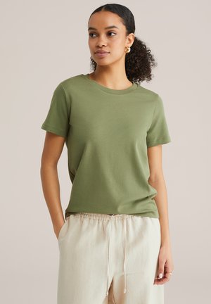 Moteris vilki alyvuogių žalia trumpomis rankovėmis marškinėlį ir šviesiai smėlio spalvos kelnės su raišteliu, turi garbanotus plaukus, surištus atgal, ir auksinius žiedinius auskarus.