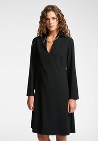Schwarzes Langarmkleid mit V-Ausschnitt und Kragen. Weicher Stoff mit lockerer Passform und dezenten Nähten. Akzentuierte silberne Kettenhalskette.