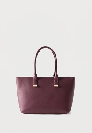 Geantă tote din piele violet închis cu două mânere și logo mic auriu al brandului pe față, pe un fundal simplu și deschis la culoare.