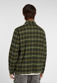 Een persoon met kort, donker haar draagt een groen geruit shirt en grijze broek, met de rug naar de camera gekeerd tegen een lichte achtergrond.