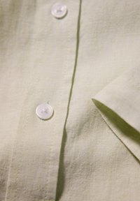 Camicetta in lino verde chiaro con chiusura a bottoni, caratterizzata da bottoni bianchi lisci, una sottile trama e un dettaglio con maniche piegate.