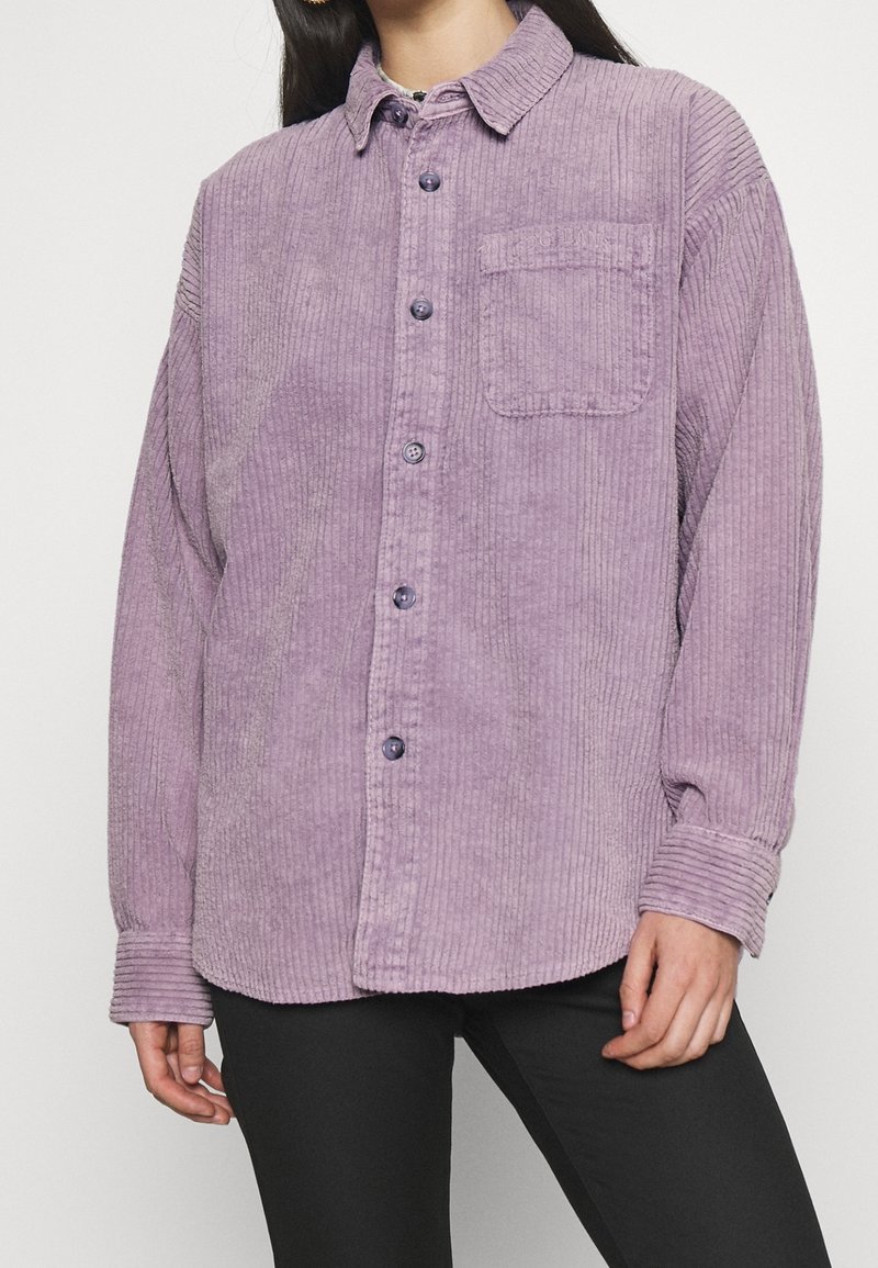 Lila kordflodsskjorta med knappslutning fram, en bröstficka och ribbad textur. Långa ärmar med uppvikta manschetter.