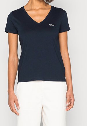 Woman wearing a navy blue V-neck short-sleeve T-shirt with a small white logo on the left chest and white pants.
