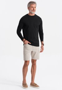Schwarzes Langarmshirt, beigefarbene Shorts und hellbraune Slipper. Das Model steht vor einem weißen Hintergrund und präsentiert ein lässiges Outfit.