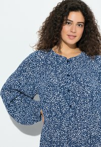 Kobieta o kręconych, ciemnych włosach, ubrana w niebiesko-białą sukienkę na guziki z długim rękawem, stojąca na tle jasnego, jednolitego tła.