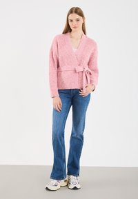 Rozā, teksturēta, apvilkšanas stila kardigāns ar sasienamo jostu, garām piedurknēm un V-veida dekoltē, kombinēts ar ziliem platām kājām biksēm un daudzkrāsainām sporta kurpēm.