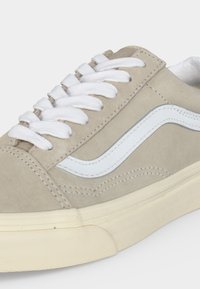 Primer plano de una zapatilla beige con cordones blancos, franja lateral blanca en forma de onda y suela de goma blanquecina sobre un fondo claro.