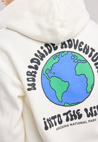 Cream-colored hoodie featuring a graphic of the Earth with green and blue colors. Text reads "WORLDWIDE ADVENTURE" and "INTO THE WILD."