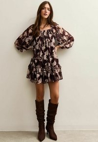 Vestido de estampado floral en marrón oscuro y crema, con mangas abullonadas y un dobladillo con volantes. Combinado con botas de tacón marrones, altas hasta la rodilla y con un diseño holgado.