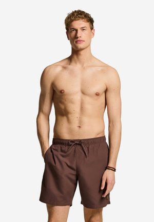 Skjorteløs mand med krøllet blondt hår iført brune badeshorts med snøre, stående med hænderne i lommerne mod en hvid baggrund.