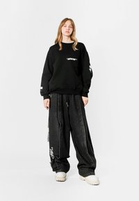 Sudadera negra con bolsillo y estampado gráfico blanco, combinada con pantalones de mezclilla negros de gran tamaño que tienen franjas laterales y cordón. Zapatillas blancas.