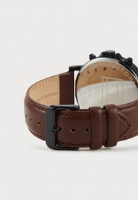 Correa de reloj de cuero marrón con una textura suave, que presenta una hebilla y acentos de metal negro. La parte trasera muestra una caja pulida y detalles grabados.