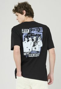 T-shirt en coton noir avec une impression graphique au dos comprenant du texte blanc et bleu ainsi que des images de quatre membres du groupe. Manches courtes, col rond.