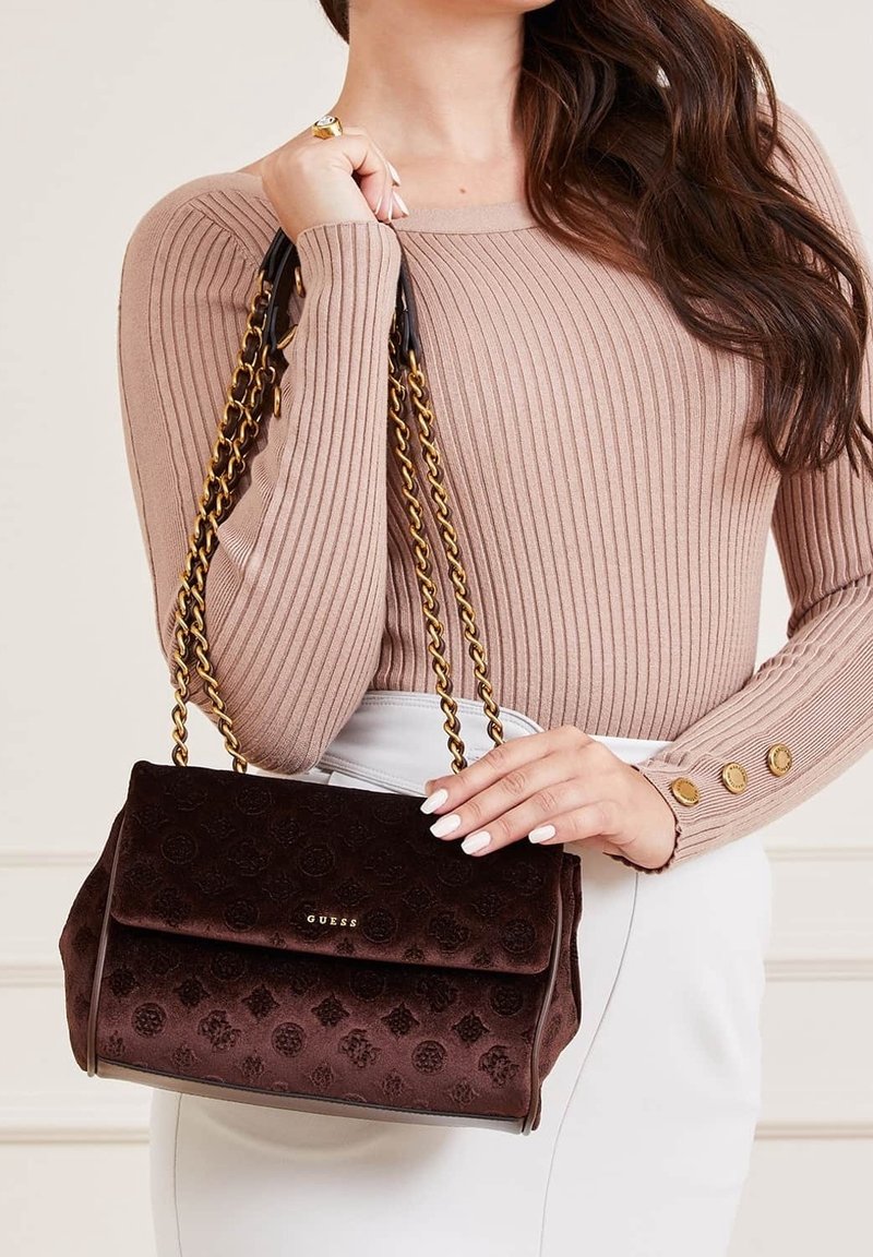 Femme tenant un sac à main Guess marron foncé avec des anses chaînes dorées, portant un pull beige côtelé et un pantalon blanc.