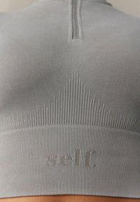 Svijetlosiva sportska majica s rebrastom teksturom, visokim izrezom i kratkim zatvaračem. Sadrži suptilno brendiranje na dnu.