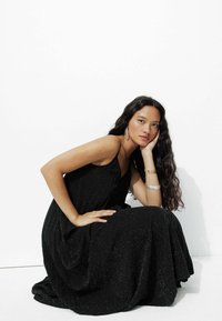 Robe noire longue au sol avec un subtil éclat, dotée de fines bretelles et d'une silhouette fluide. Modèle assis avec des cheveux ondulés et des bijoux minimalistes.