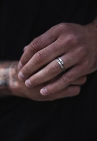 Due anelli d'argento impilati su una mano, uno con una finitura liscia e l'altro con una superficie texturizzata e incisioni. Sulla mano sono visibili dei tatuaggi.
