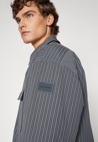 Randi grå skjorte laget av lett stoff. Har krage, brystlomme og et Calvin Klein-merke på skulderen.