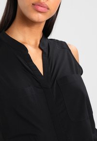 Blusa nera senza maniche con scollatura a V, due tasche sul petto e una texture liscia con una leggera lucentezza.