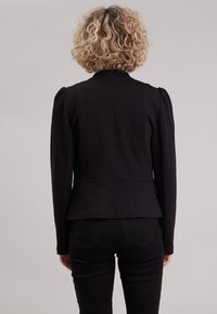 Blazer noir ajusté avec épaules rembourrées, taille cintrée et design peplum. Confectionné en tissu lisse, avec un détail de couture simple au dos.