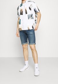 Camisa blanca de manga corta con impresiones fotográficas coloridas, combinada con shorts de mezclilla azul, puños enrollados y zapatillas blancas con calcetines.