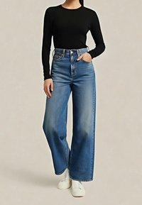 Femme portant un haut noir à manches longues, un jean bleu taille haute à jambes larges, des baskets blanches, avec une main dans la poche, sur fond uni.