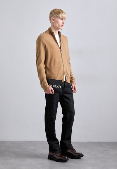 Tânăr îmbrăcat cu un pulover camel tip zip-up, pantaloni negri și pantofi maro, ținând un clutch de marcă Emporio de culoare neagră, stând pe un podea gri.