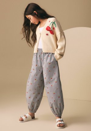 Young girl wearing cream cardigan with cherry design, striped pants with small red fruit prints, and white sandals, standing with hands in pockets.