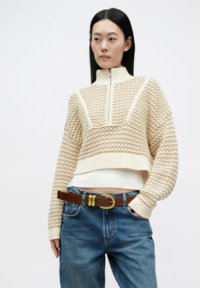 Pulover tricotat în culori bej deschis și crem, cu guler înalt, fermoar pe jumătate și design scurt; asortat cu blugi albaștri și o curea maro.