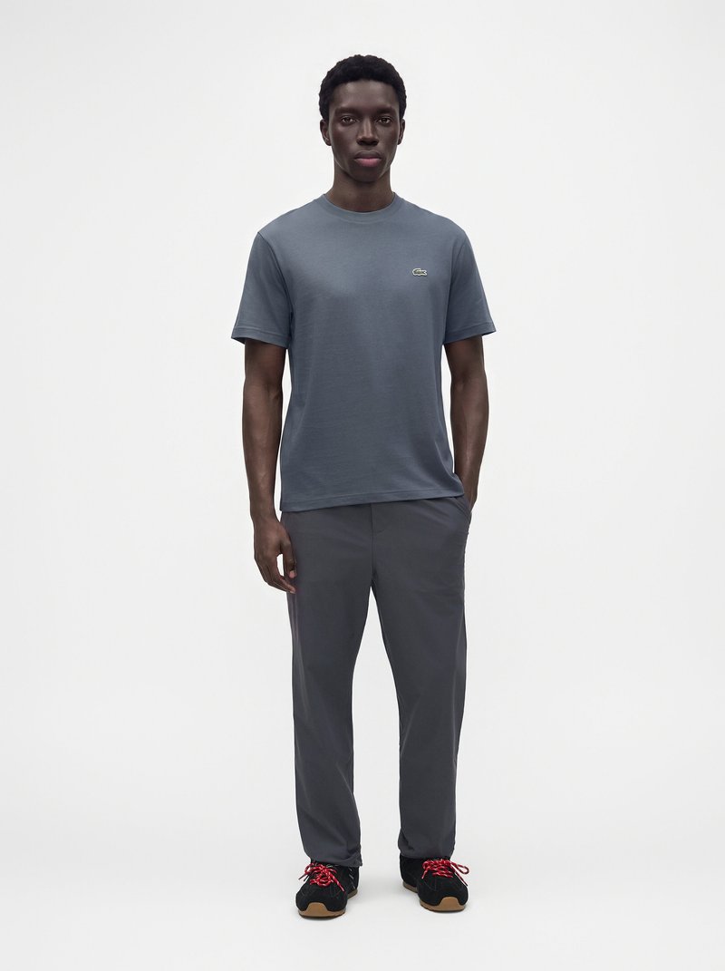 Homme debout portant un t-shirt gris à manches courtes, un pantalon gris foncé et des baskets noires avec des lacets rouges sur un fond blanc uni.