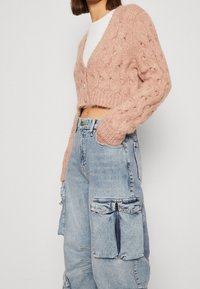 Růžový pletený zkrácený kardigan s texturovaným vzorem, dlouhými rukávy a třemi knoflíky. V kombinaci s vybledlými modrými cargo džínami.